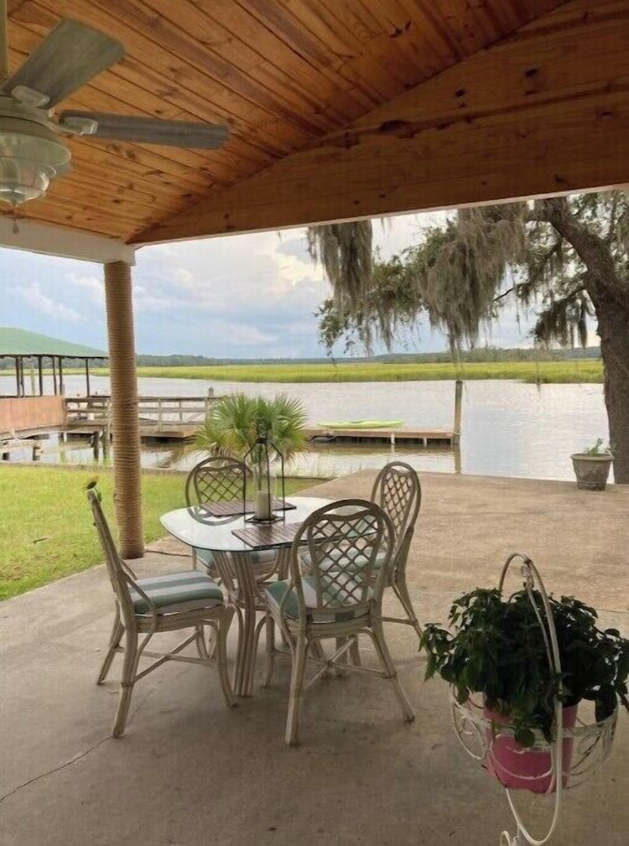 A covered patio with a table and chairs, overlooking a body of water and a grassy landscape.