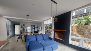 Una sala de estar moderna con un sofá seccional azul, una zona de comedor con mesa y sillas, una cocina al fondo y una chimenea de piedra con vistas al exterior.