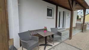 A covered patio area with a table and two chairs, a bench, and a potted plant.