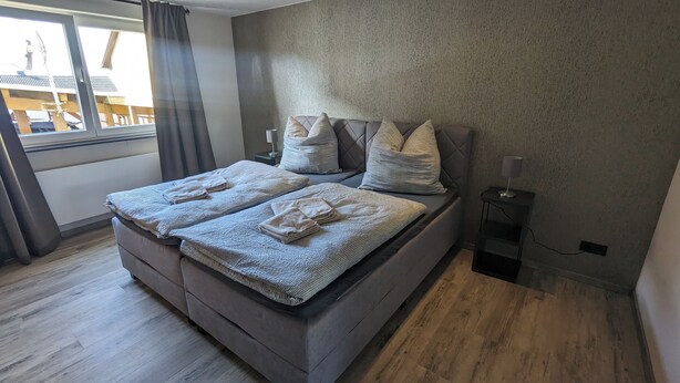 A double bed with a grey headboard, two pillows, and folded towels. There is a bedside table with a lamp, a window with curtains, and a view of a building outside.