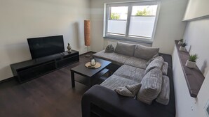 A modern living room with a large flat-screen TV, a grey sofa, a coffee table, and a window with blinds.