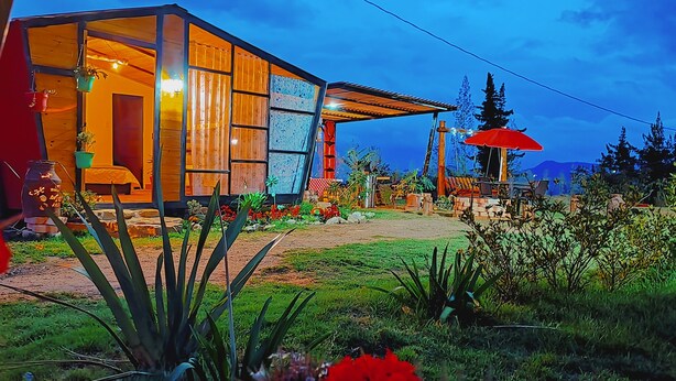 A modern house with large windows, a covered porch, and a garden with various plants and flowers. - LA TATA CABAÑA  ZON DE CAMPING  (Cucunubá)