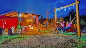 A rustic outdoor setting with a covered seating area, a swing set, and a red building with a corrugated metal roof. - LA TATA CABAÑA  ZON DE CAMPING  (Cucunubá)