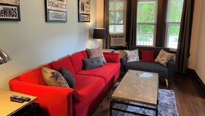 Un salon avec un canapé rouge, un canapé foncé et une table basse. Il y a deux fenêtres avec des rideaux, un climatiseur et une lampe sur une table d’appoint.