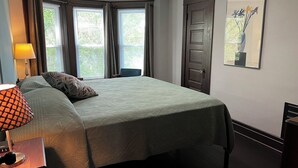 Un lit bien fait, avec une couvre-lit verte et un coussin à motifs, une lampe de chevet, une œuvre d’art encadrée au mur et une porte avec une fenêtre.