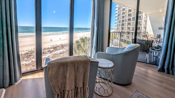 Una habitación con un amplio ventanal que da a la playa, un balcón con mobiliario exterior y vistas a un edificio de gran altura.