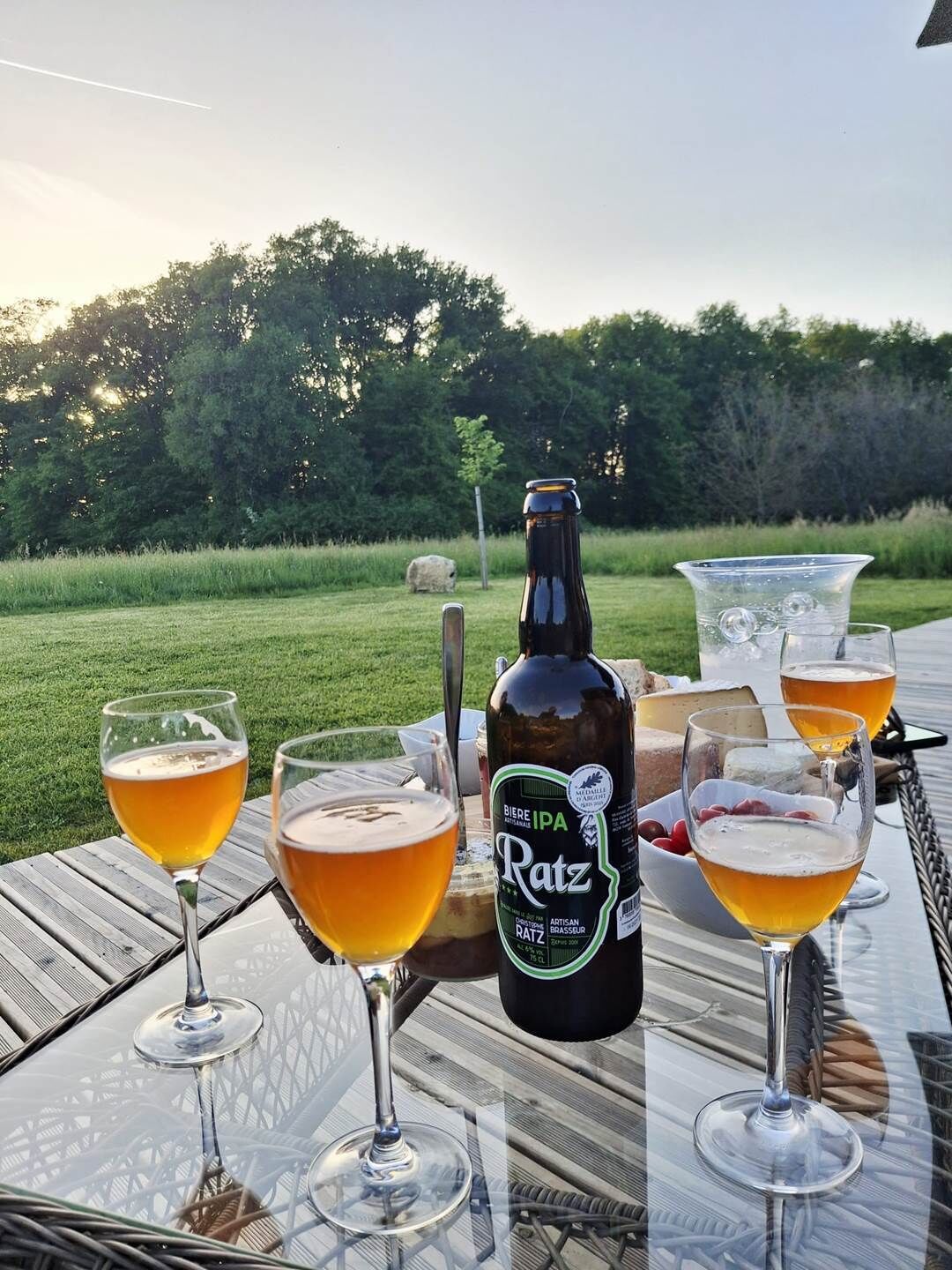 A bottle of beer, glasses of beer, and a bowl of grapes on an outdoor table.