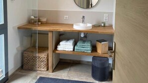 A modern bathroom with a glass shower enclosure, a wooden vanity with a round mirror, and a black trash bin.