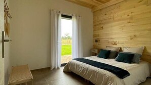 A bedroom with a wooden ceiling, a bed with white bedding and blue pillows, a small stool, and a view of the outdoors through a window with white curtains.