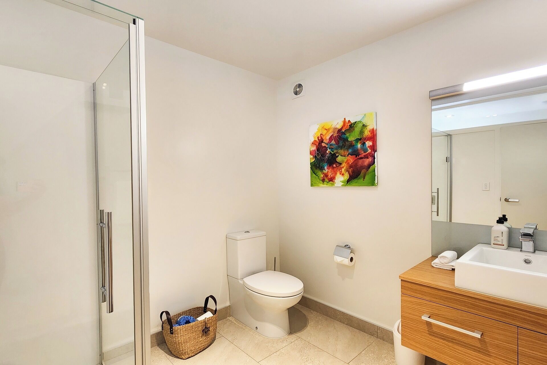 Une salle de bain avec une douche à parois de verre, des toilettes, un lavabo avec un miroir et une peinture abstraite aux couleurs vives accrochée au mur.