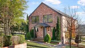 A two-story brick house with a gabled roof, a front porch, and a well-maintained garden with hedges and trees.