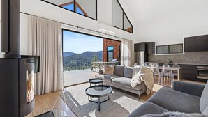A modern living room with a sofa, coffee table, and TV. It features large windows with a view of the outdoors.