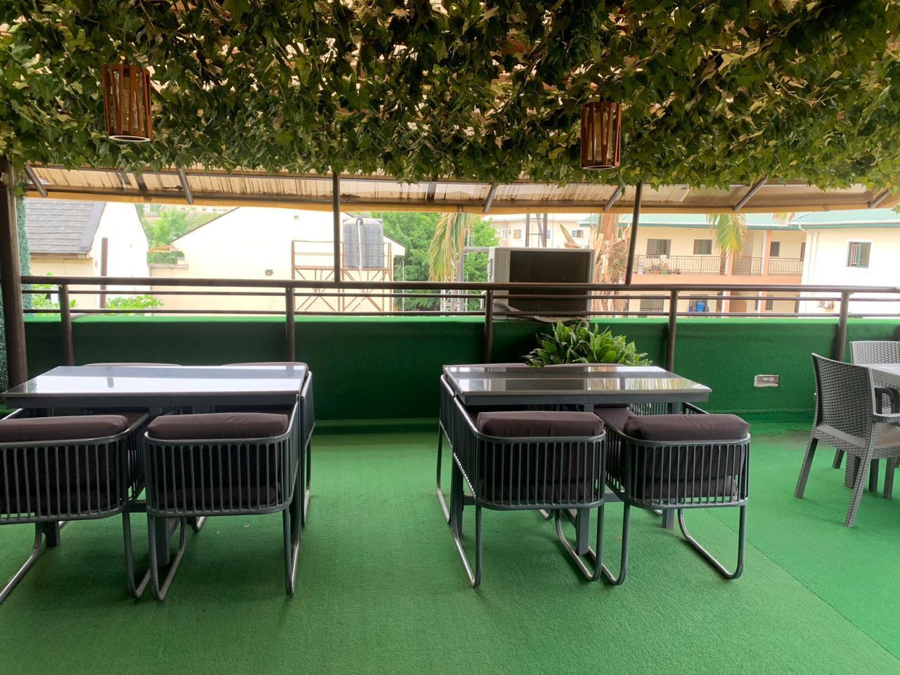 An outdoor patio area with green artificial turf, metal tables and chairs, hanging lanterns, and a view of buildings in the background.