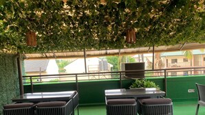 An outdoor seating area with tables and chairs, covered by a green roof, and a view of buildings in the background.