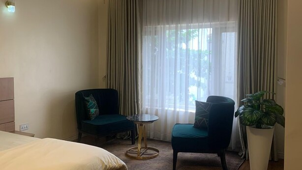 A hotel room with a bed, two chairs, a small table, a potted plant, and a large window with curtains.