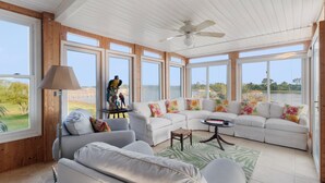 A spacious living room with a white sofa, a coffee table, and a lamp. Large windows offer views of a lake and greenery.