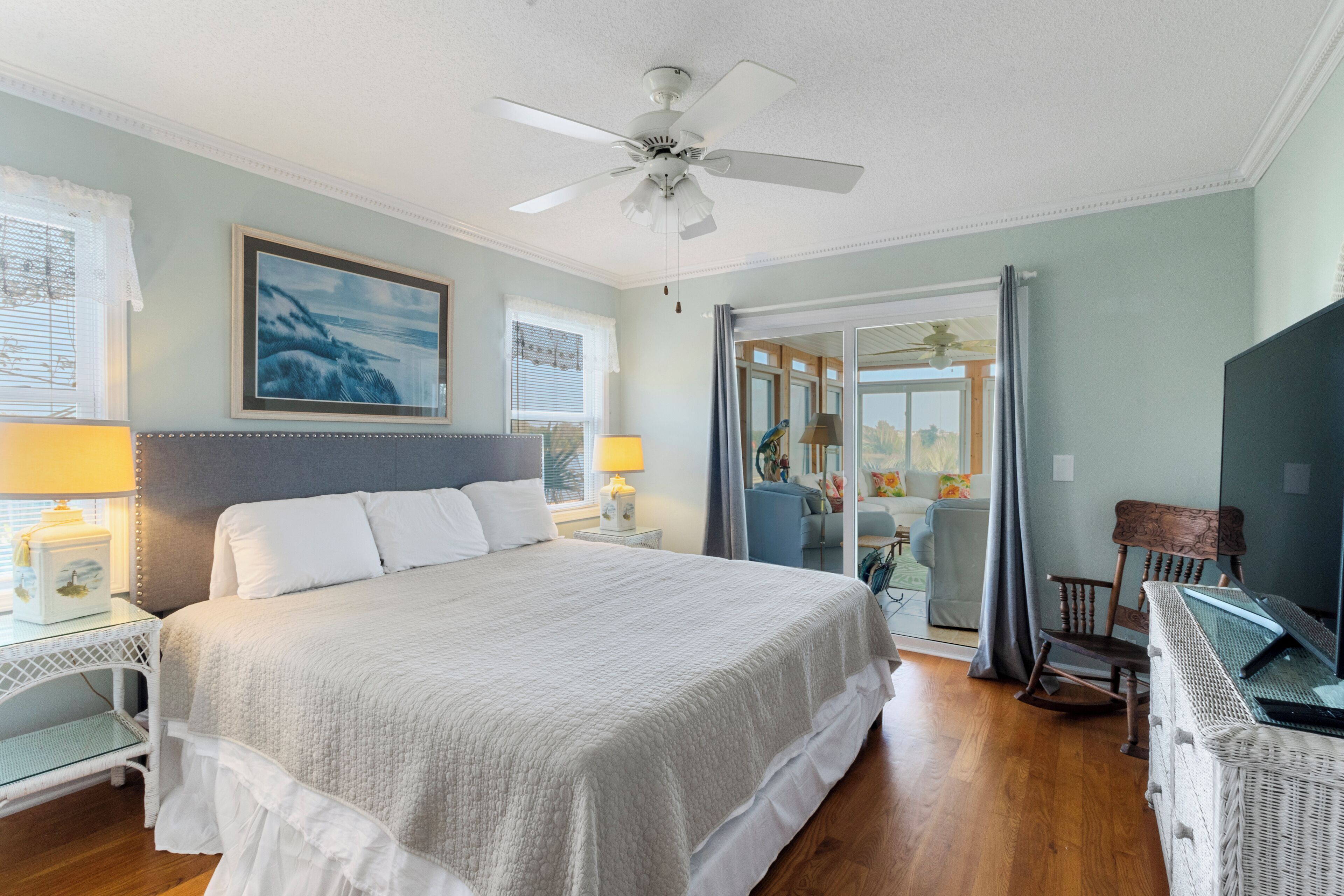 A bedroom with a large bed, two bedside tables with lamps, a ceiling fan, and a view to the outside.