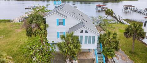 A white house with blue shutters, surrounded by a grassy yard and palm trees, with docks extending into a body of water.