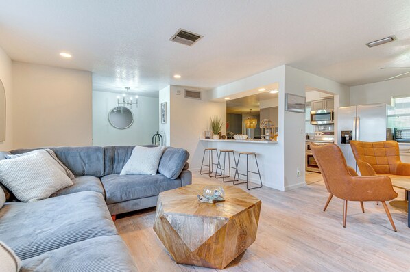 Ein modernes Wohnzimmer mit einer Eckcouch, einem Holztisch und einer Küchenzeile mit Barhocker und Kühlschrank.