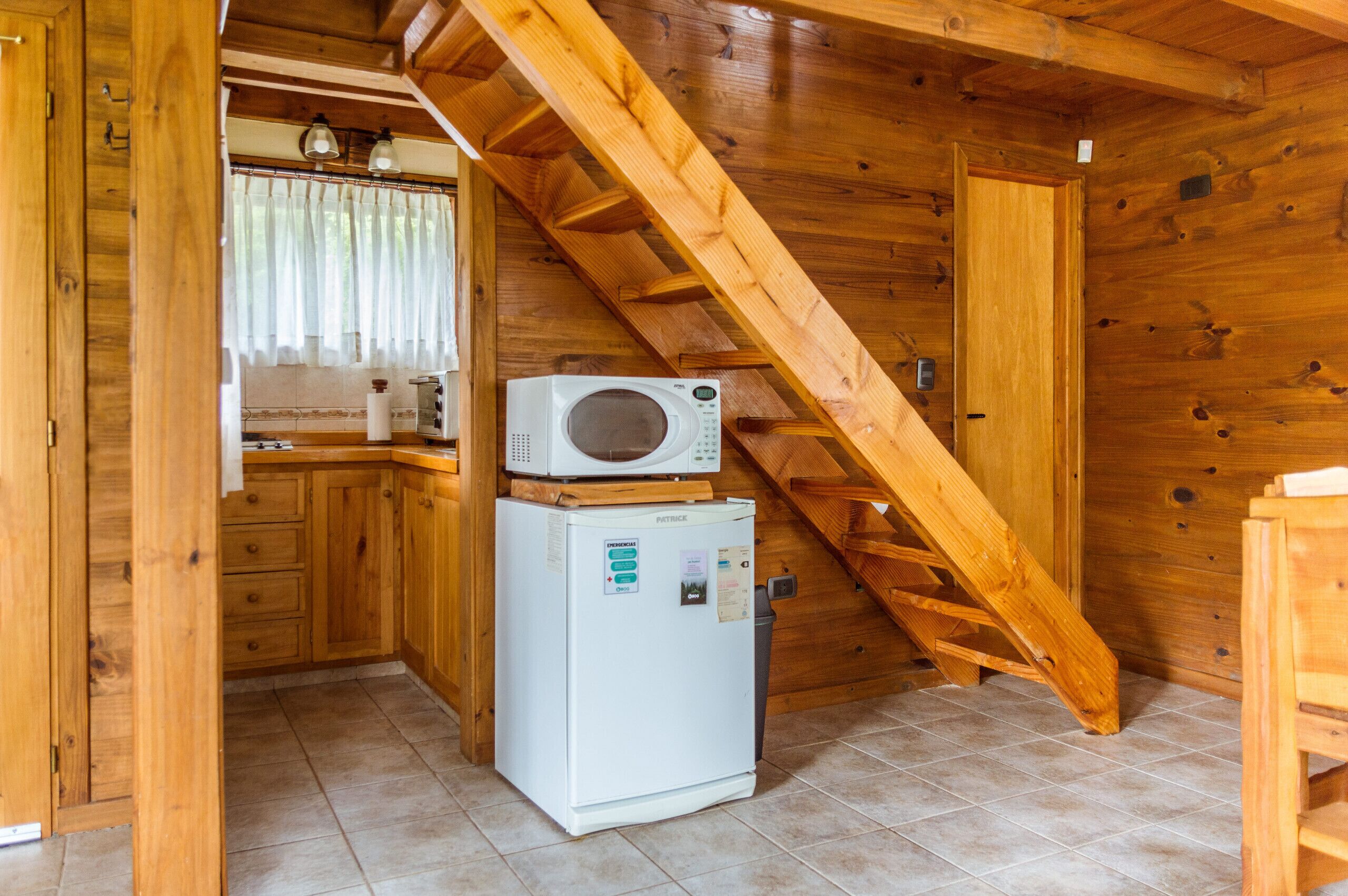 Une cuisine compacte dotée de meubles en bois, d’un four à micro-ondes et d’un réfrigérateur. Un escalier en bois mène à l’étage supérieur.