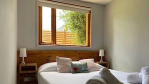 Un dormitorio con cabecera de madera, dos mesitas de noche con lámparas, una cama con ropa blanca y almohadas decorativas, una ventana con vistas a follaje y una toalla enrollada sobre la cama.