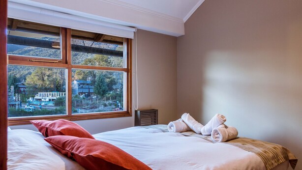 Ein Hotelzimmer mit einem großen Fenster, einem Bett mit weißen Leinentüchern, zwei roten Kissen und einem aufgerollten Handtuch auf dem Bett.