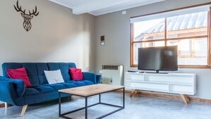 Ein Wohnzimmer mit einem blauen Sofa, einem hölzernen Couchtisch, einem aufgehängten Hirschkopf, einem großen Fernseher und einem Fenster mit Blick auf ein Gebäude.