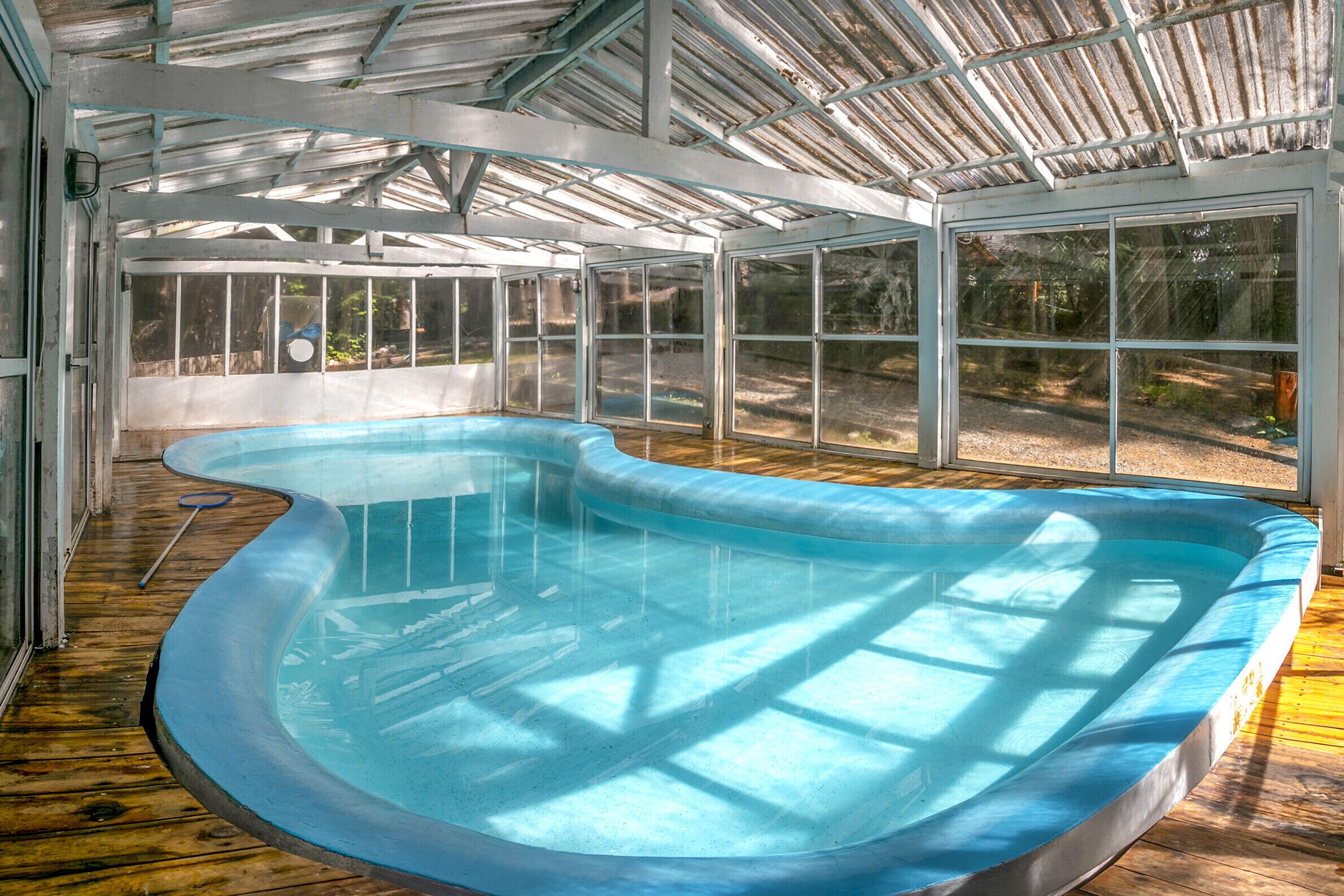En indendørs swimmingpool med trægulv og glasvægge, omgivet af en metal konstruktion.