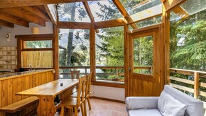 Una cabaña de madera con comedor, sofá y ventanas amplias que dan a un bosque.
