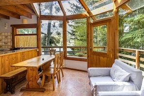 A wooden cabin with a dining area, a sofa, and large windows offering a forest view.