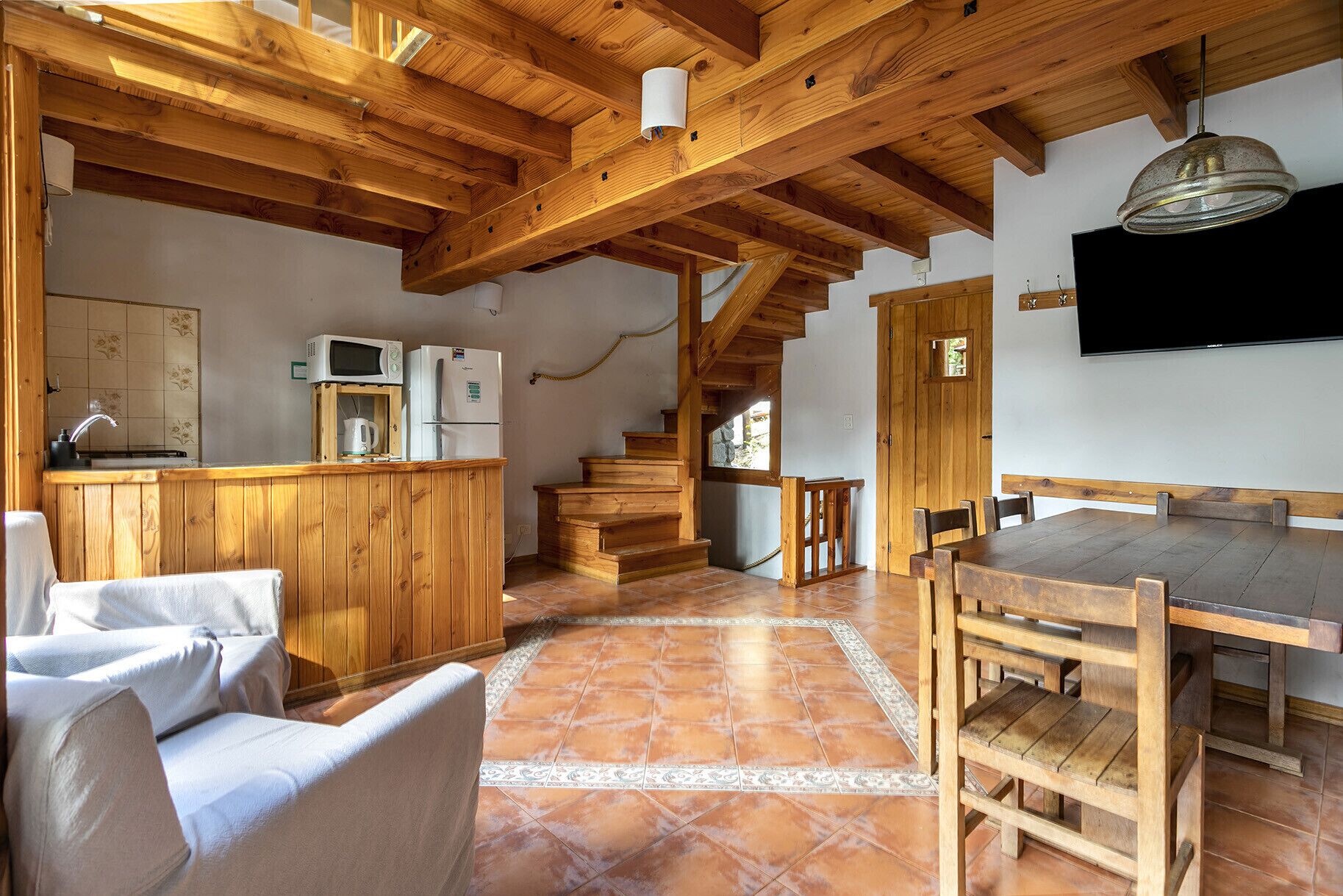 Un interior rústico con vigas de madera, una zona de cocina con electrodomésticos, una mesa de comedor y una escalera.