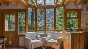 Una cabaña acogedora con dos sillones mullidos, una mesa de madera y una zona de cocina con lavabo. Amplias ventanas ofrecen vistas a una zona montañosa boscosa.