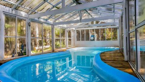 Una amplia piscina cubierta con un deck de madera que la rodea, dentro de una estructura de vidrio y metal.