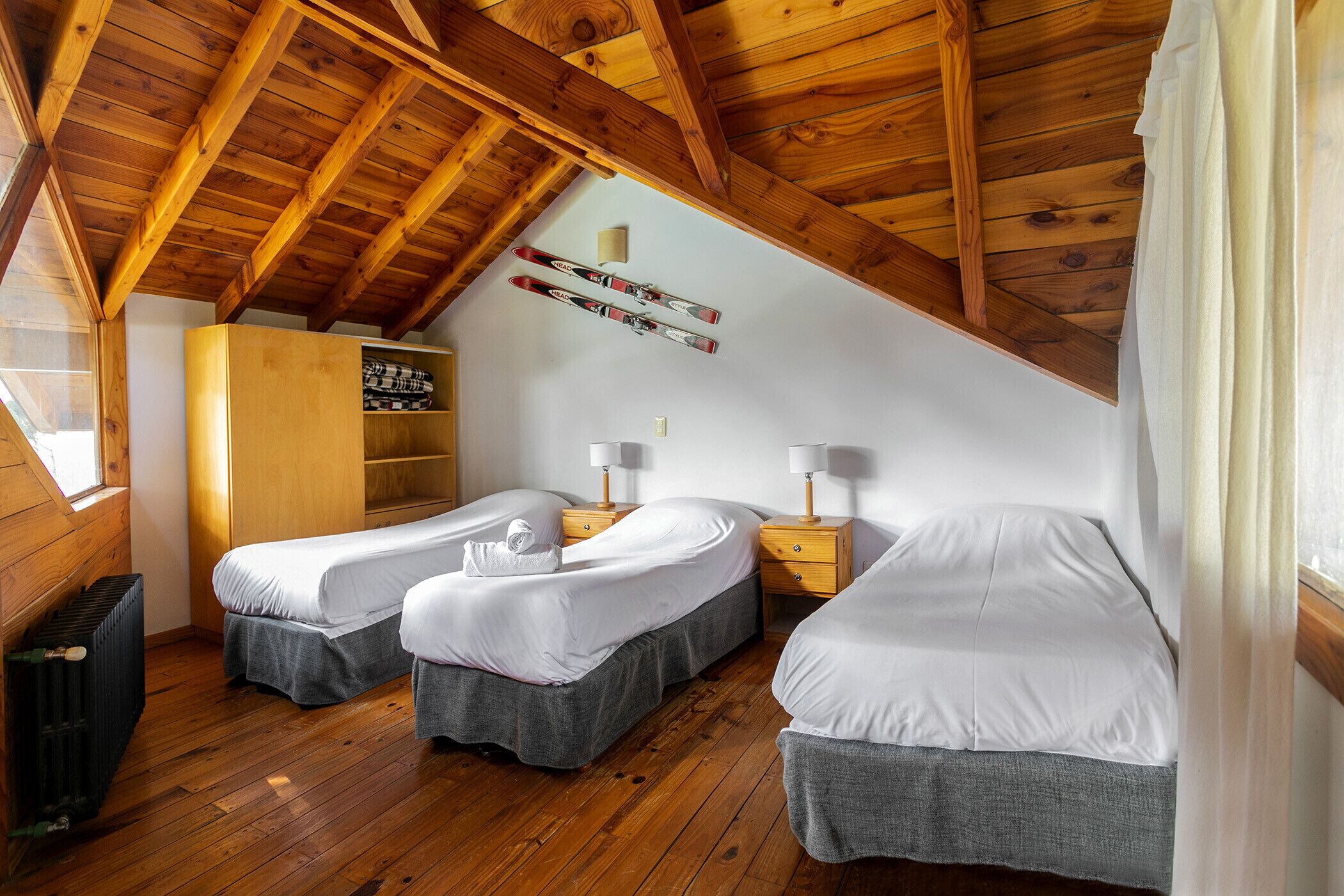 Ein gemütliches Schlafzimmer im Dachgeschoss mit Holzbalken, zwei Betten mit grauer Bettwäsche, einem Holzschrank und einem Skiständer an der Decke.