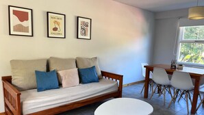 A modern living room with a wooden sofa, a white coffee table, and a dining area with a wooden table and chairs. There are three framed artworks on the wall.