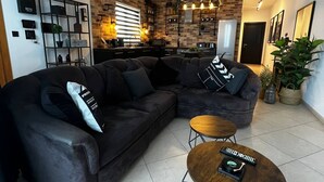 A modern living room with a sectional sofa, a coffee table, and a kitchen area with a brick wall and pendant lighting. - Trendy Seaside Escape |Designer Finished Penthouse

 (Wied il-Ghajn)