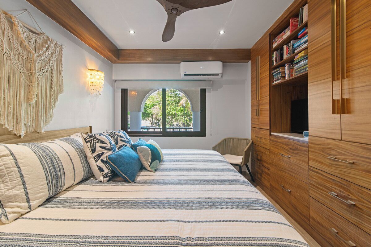 Ein Schlafzimmer mit einem Bett, einem Holzschrank, Wandkunst aus Makramee und einem Fenster mit Blick ins Grüne.