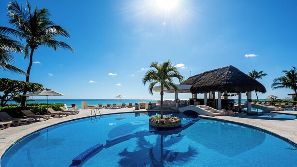 A pool with lounge chairs and umbrellas, surrounded by palm trees and a view of the ocean.