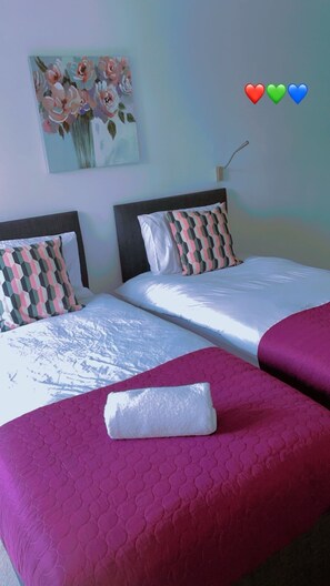 Ein Hotelzimmer mit zwei Betten, jedes mit einem gemusterten Kopfkissen und einer weißen Tagesdecke. Darüber hängt ein Gemälde an der Wand und ein Nachtlicht steht daneben.