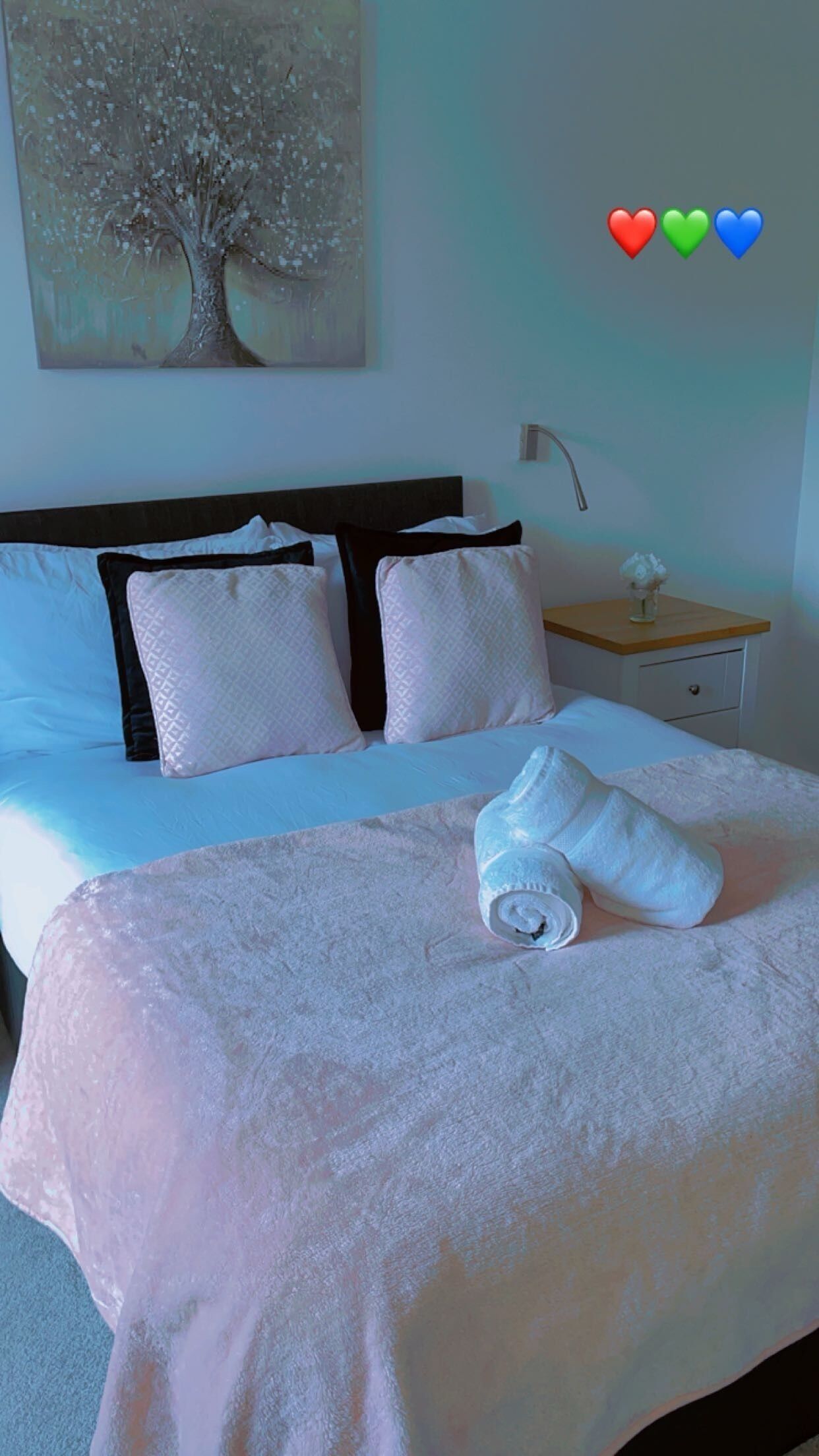 A neatly made bed with a white duvet and pillows, a wooden nightstand with a lamp, and a painting of a tree on the wall.