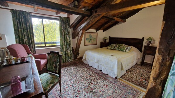 A bedroom with a bed, a chair, a table with items, a window, and a wooden ceiling.
