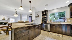 A modern kitchen with dark wood cabinets, a large island, pendant lighting, and a view of the outdoors through a large window. - Cozy Fall Retreat w/ Swim Spa (Tigard)