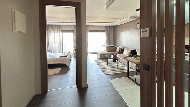 Ein modernes Hotelzimmer mit einem großen Bett, einer Sitzecke, einem Schreibtisch mit einem Laptop und Blick auf die Stadt durch große Fenster.