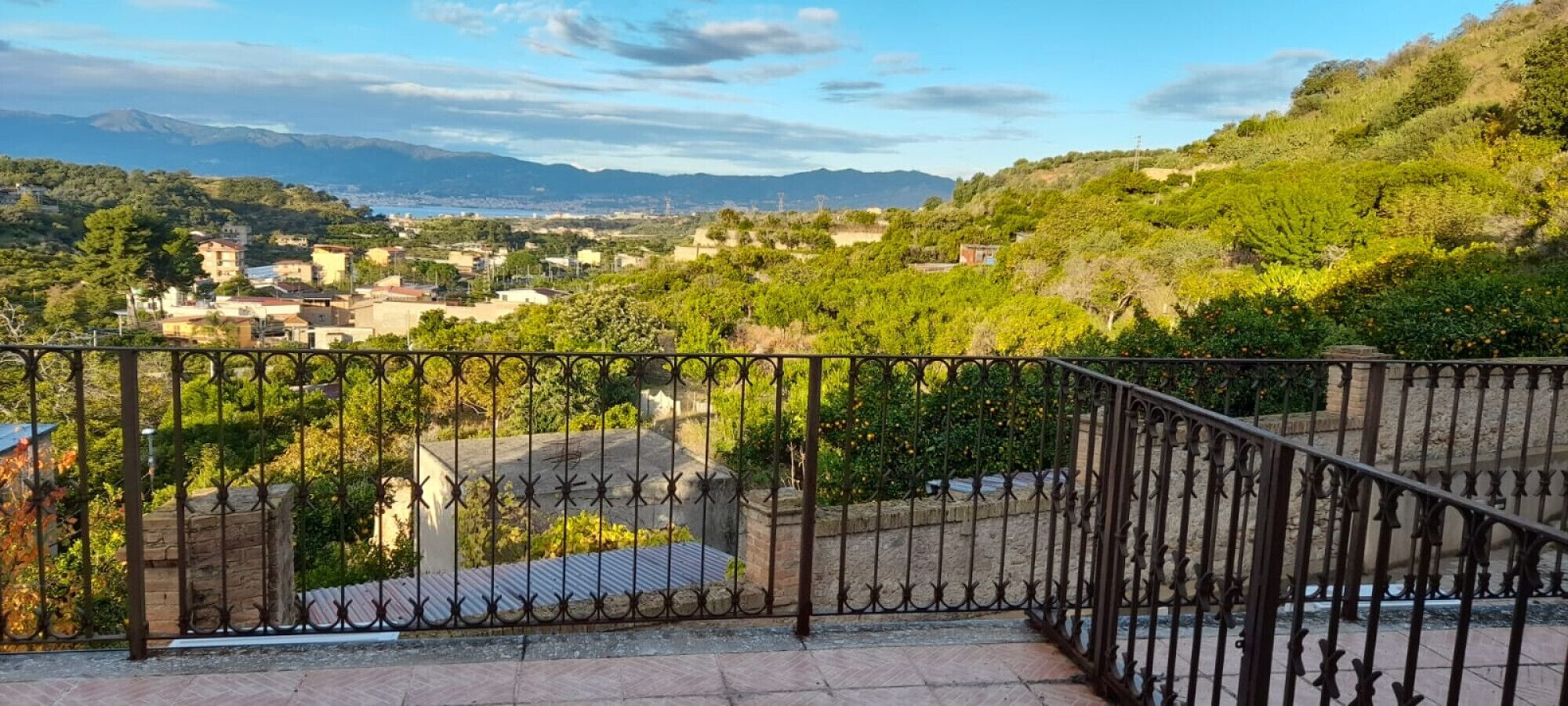 Un balcon doté d’une balustrade en fer forgé, donnant sur une ville avec ses bâtiments et sa verdure.