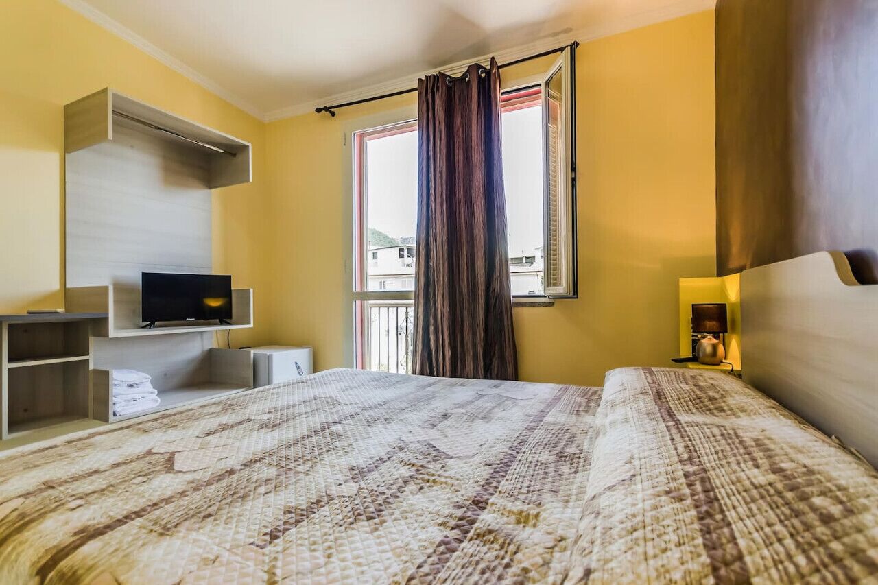 A hotel room with a bed, a TV, a shelf with towels, a window with curtains, and a nightstand with a lamp.