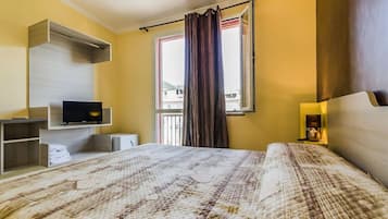 A hotel room with a bed, a TV, a shelf with towels, a window with curtains, and a nightstand with a lamp.