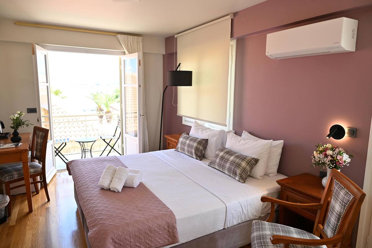 A hotel room with a bed, bedside tables, a desk, a chair, a balcony with a table and chairs, and an air conditioning unit.