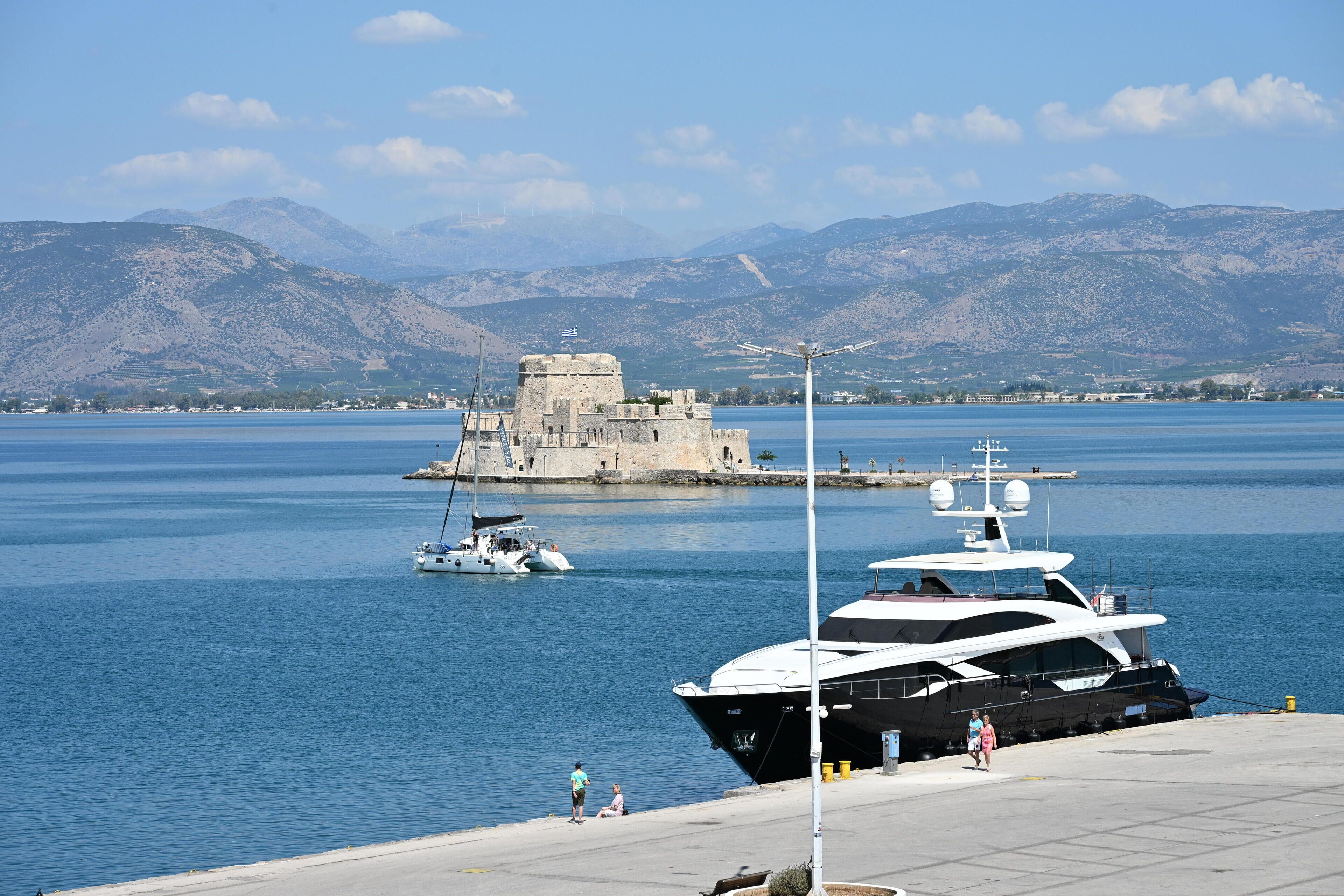 Uma marina com iates atracados, um castelo ao fundo e montanhas à distância.