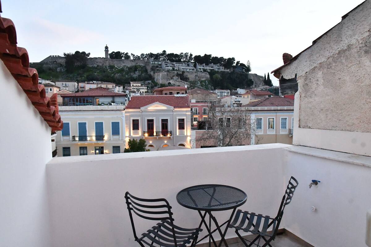 A balcony with two chairs and a table, overlooking a cityscape with historic buildings and a hill with a tower.