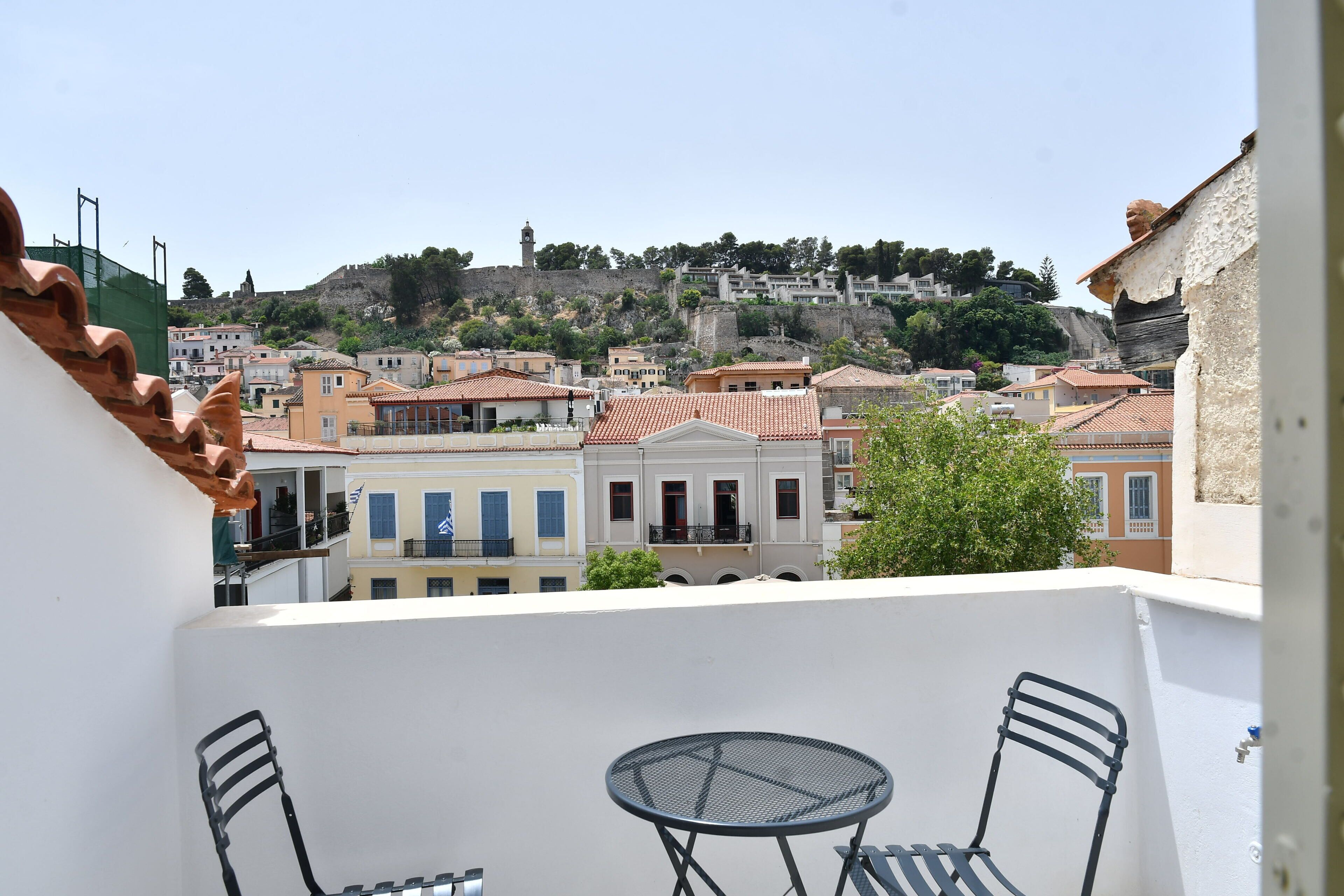 Un balcón con mesa y sillas, con vistas a un pueblo de edificios coloridos y una colina con una torre.
